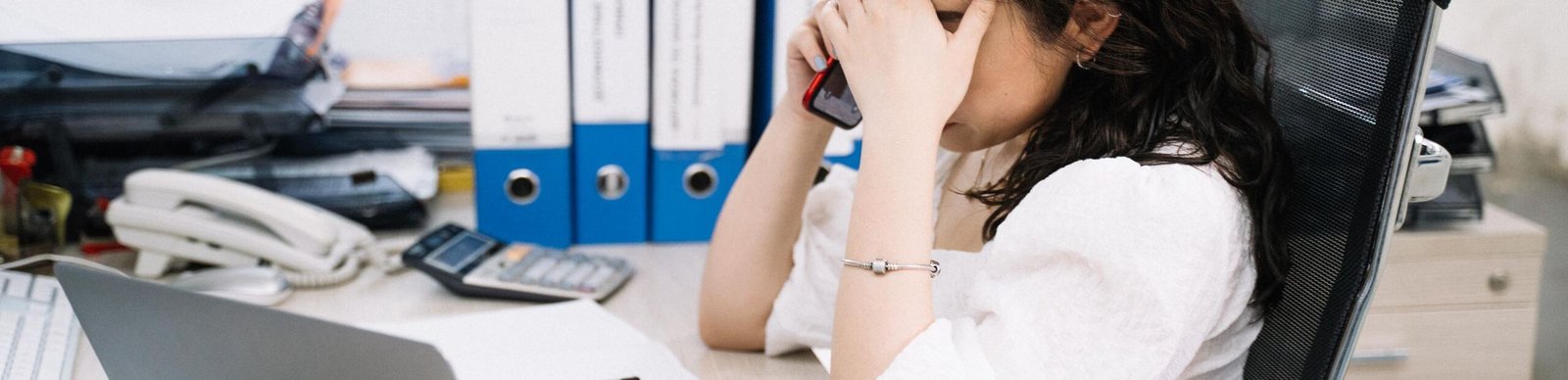A woman in an office sitting at a desk, visibly stressed, using a phone. Workplace stress concept.
